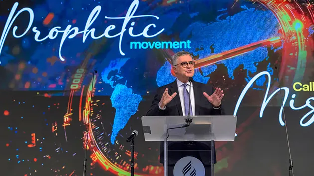 Мужчина в костюме выступает за кафедрой на фоне экрана с надписью «Prophetic movement»