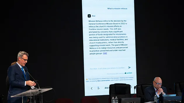Чоловік за трибуною виступає на тлі великого екрана з текстом чат-бота про місію церкви