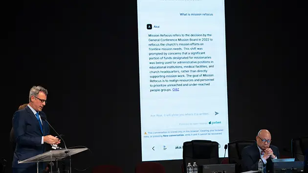 Чоловік за трибуною виступає на тлі великого екрана з текстом чат-бота про місію церкви