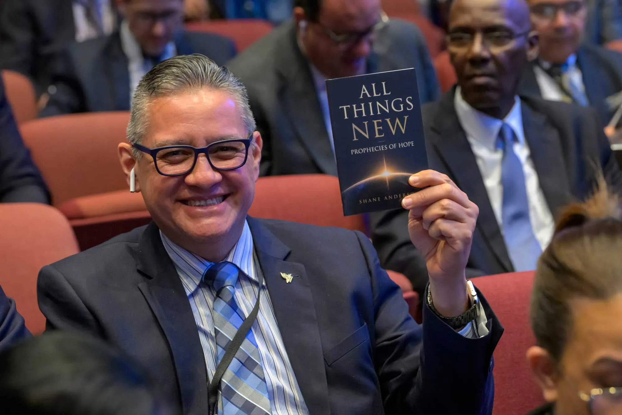 Чоловік у залі засідань усміхнено тримає в руці книгу «All Things New»