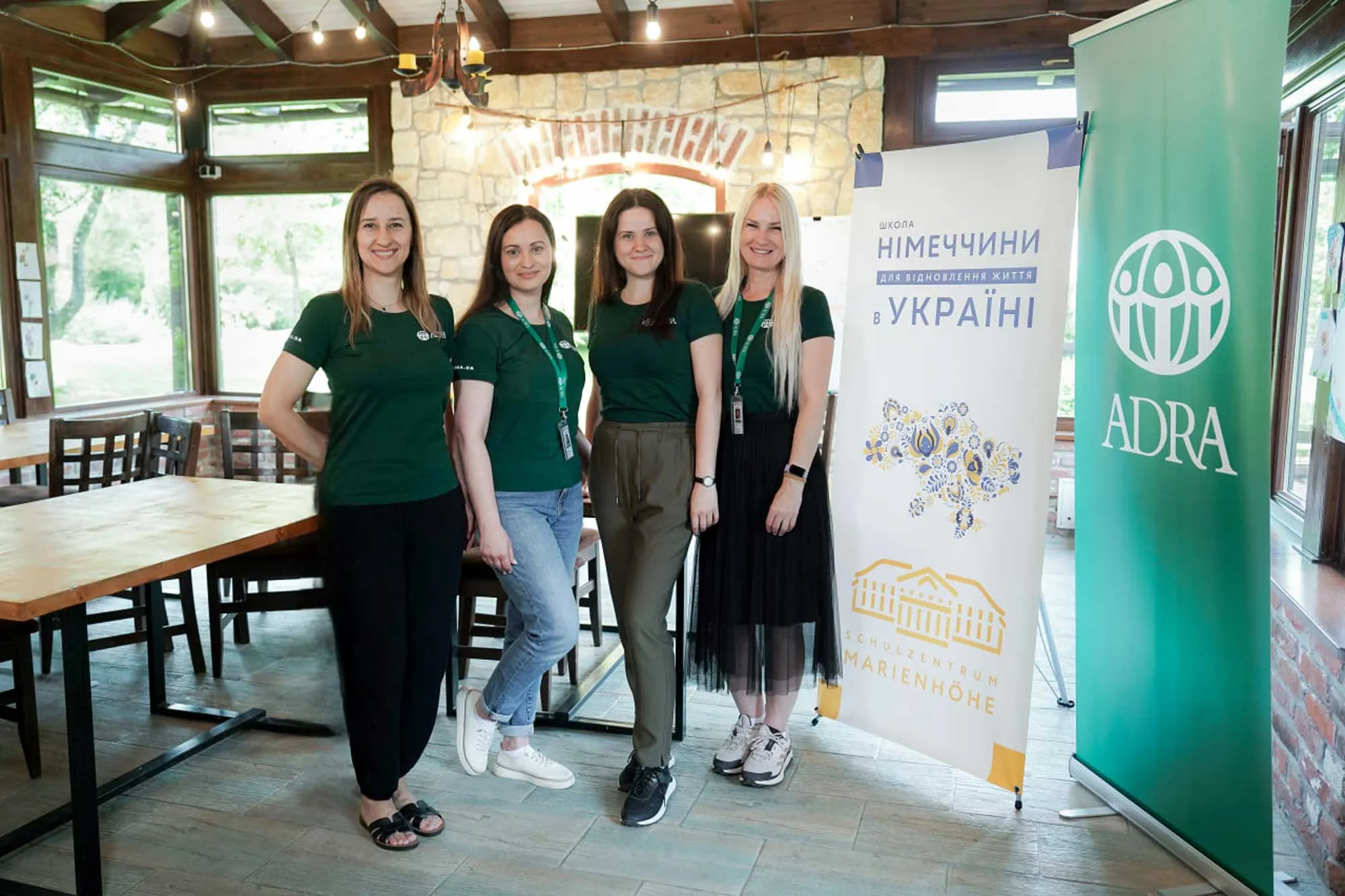 Чотири жінки в зелених футболках стоять біля банерів з логотипом ADRA та написом «Школа Німеччини для відновлення життя в Україні»
