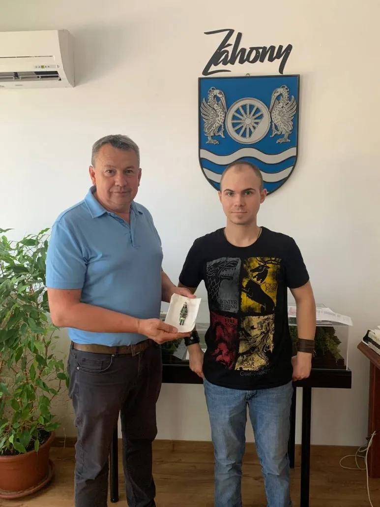 Двоє чоловіків стоять під гербом Захоня, один тримає невелику пластилінову фігурку людини