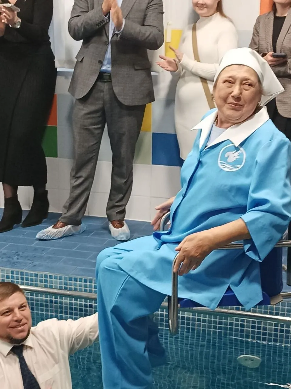 Жінка в блакитному хрестильному одязі біля краю басейну, поруч чоловік допомагає їй спуститися у воду