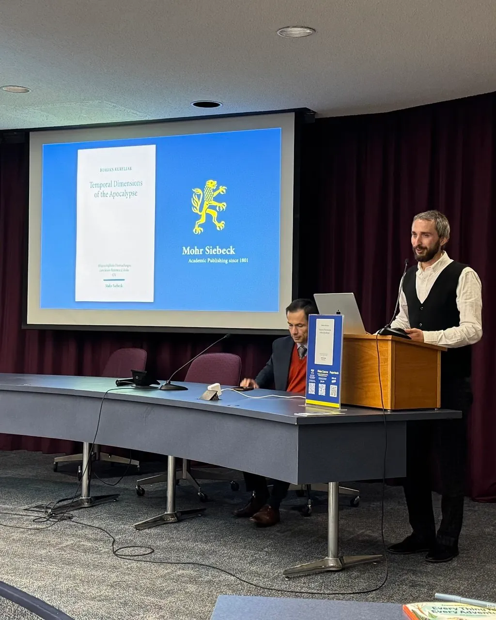 Чоловік за трибуною виступає на тлі екрана з обкладинкою книги «Temporal Dimensions of the Apocalypse»