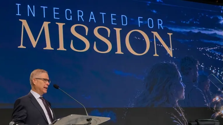 Чоловік у костюмі виступає з трибуни на тлі екрана з написом «Integrated for Mission»