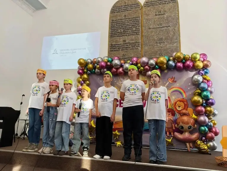 Дети в одинаковых футболках и цветных шапочках поют на сцене с микрофонами