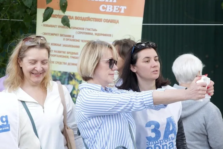 Женщины фотографируются на смартфон на фоне стенда о пользе солнечного света