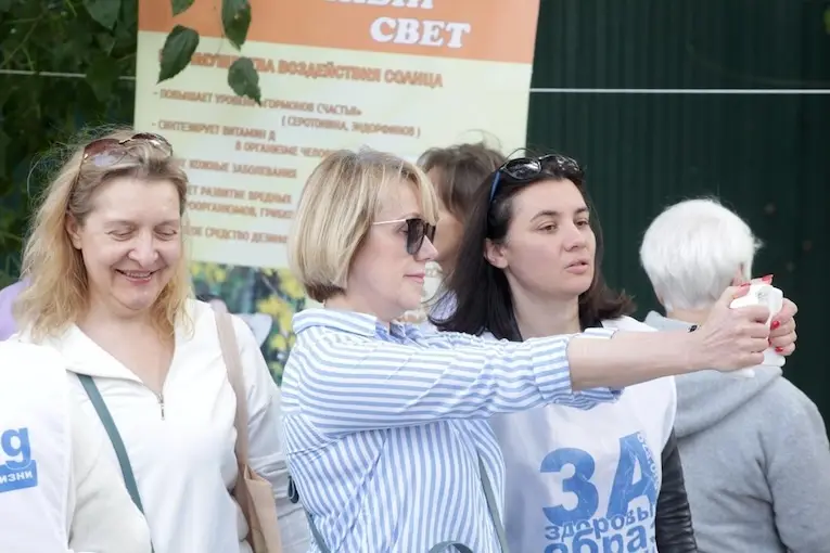 Женщины фотографируются на смартфон на фоне стенда о пользе солнечного света